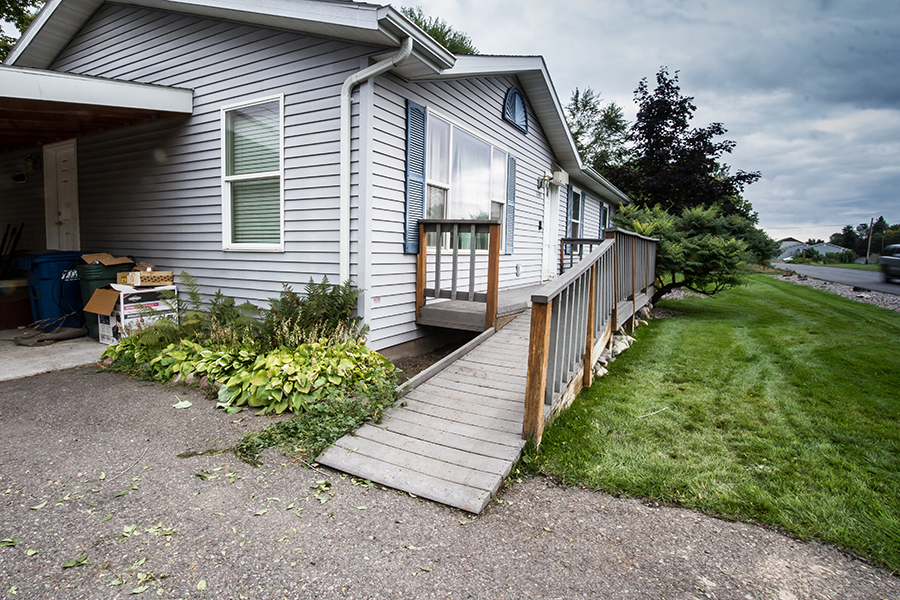 The grey ramp in the front of the house hangs nearly a foot into the drive. Several light green bushes press against the side with a safety guard to prevent wheels from sliding off. On the street-facing side, there is a handrail that ends just before the driveway.